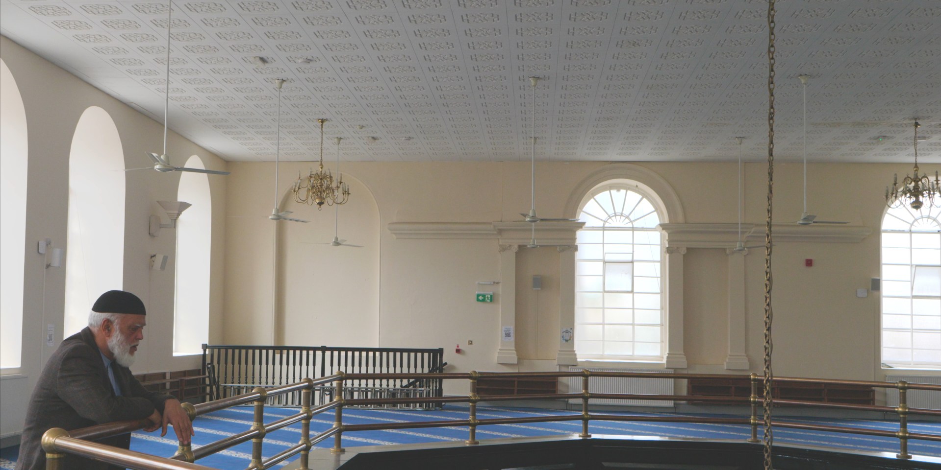 An elderly man leans on a brass rail in the gallery of a mosque, looking out over the blue-carpeted prayer hall and its arched windows.